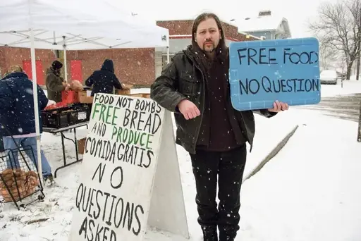 Middle-aged man standing in the snow near a Food not Bombs food distribution site with a sign saying "Free food, no question".