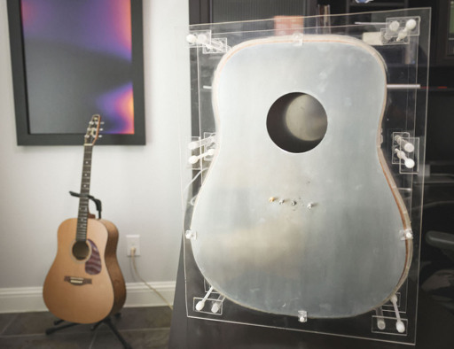 The image shows a photonic guitar in the foreground. The photonic guitar is shaped like an acoustic guitar but is made out of pure aluminum, not wood. Further, while an acoustic guitar has bracing internal to the instrument the photonic guitar uses acrylic as an exoskeleton.