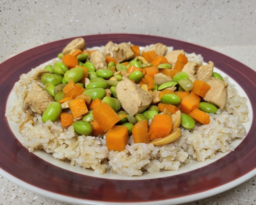 a plate of bourbon chicken made of chicken, carrots, edamame, and cashews served over brown rice